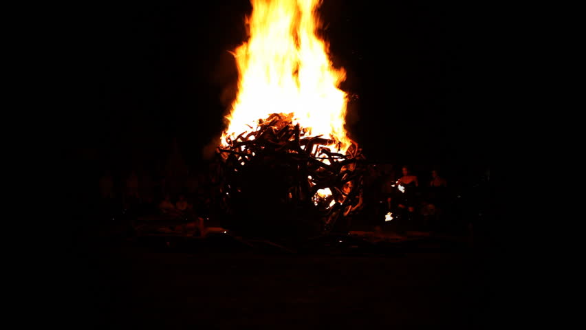 A huge sphere of wood is lt up and spreading high flames in front of black