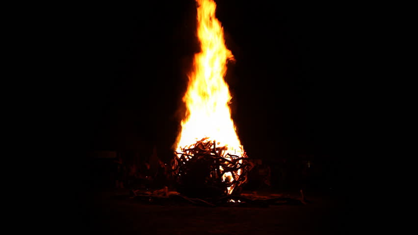 A huge sphere of wood is lt up and spreading high flames in front of black