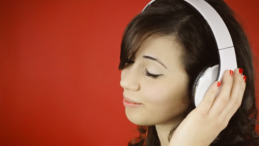 A pretty young woman listening to music with white headphones, looking careless. Close-up on red background, room for copyspace.
