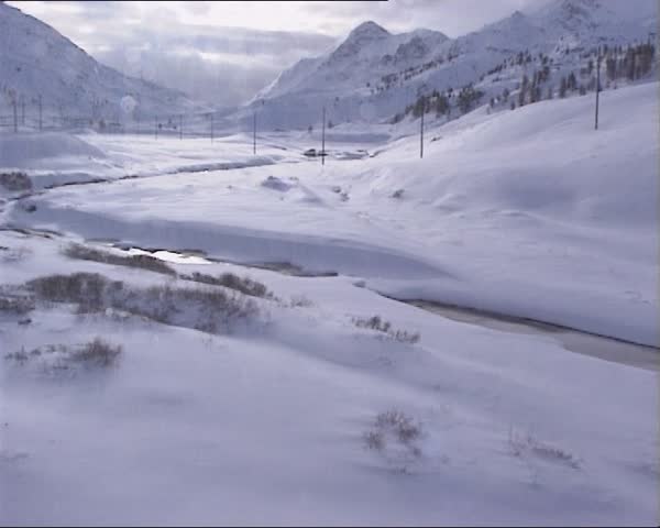BERNINABAHN, SWITZERLAND - WINTER 1995: vehicle shot from riding train at snow covered valley and stream. Blowing snow. Bernina Railway, Switzerland, added to the list of UNESCO World Heritage Sites.