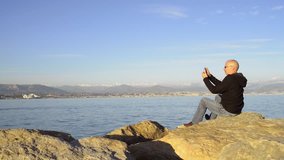 Mobile photography/tourism: mature man sat on rocks uses smart phone to shoot video or panoramic photo of the Mediterranean sea and Alps. Sunny evening, snow on mountains. Antibes, France