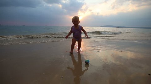 blonde baby girl in purple swimsuit play with ball in the waves at sunset