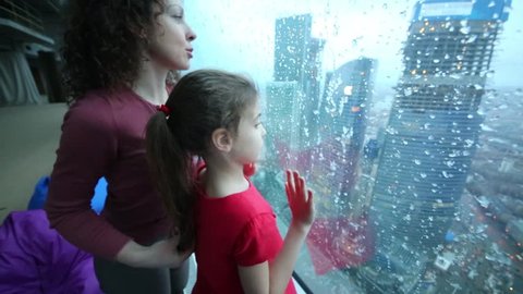 Mother and daughter look through large window with frozen drops on modern city