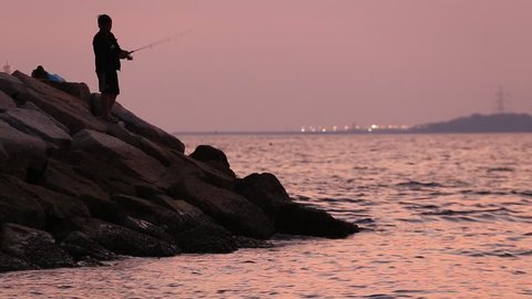  A man fishing from the breakwaters wall with industrial background during sunset, Thailand