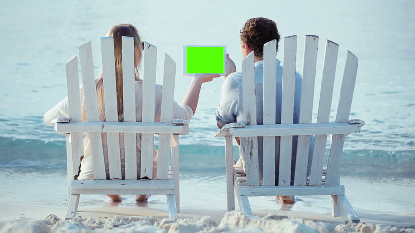 Young couple sitting back in the wooden chaise-longues on the beach. They holding tablet computer with green screen and looking at it