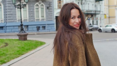 Young, beautiful, attractive woman in a stylish coat walks through the city streets. She laughs, poses for the camera, her hair blowing in the wind, looking very happy. slow motion