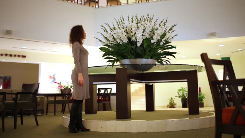 Young woman stands in hall near to big flowerpot with flowers, then leaves room.