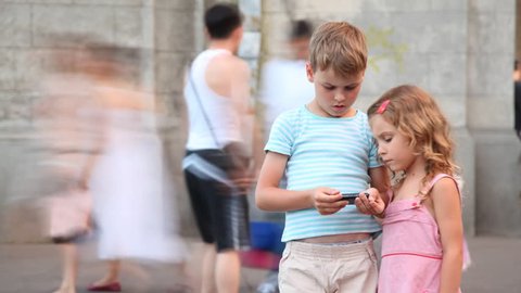 The boy plays on a hand-held device, the girl observes of it, people move behind them. Time lapse. 