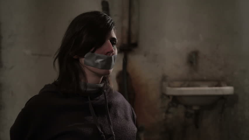 Beaten female hostage sits tied in dirty basement, kidnapper coming and put adhesive tape on her mouth. Abducted girl. Gagged woman. Violence against women. Victim of kidnapping and human trafficking. | Shutterstock HD Video #8696398