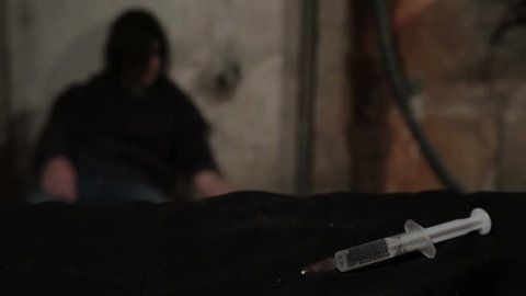 Syringe with heroine close up. Junkie female have crisis, waiting for the dose of heroin. Guy takes heroine dose and injecting in vein of girl who sits in corner. Syringe in focus, drugging blurred.