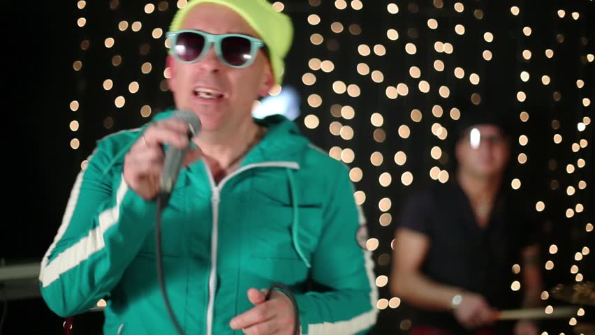Singer stands at microphone in studio with drum set.
