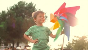 At sunset in summer day happy child wearing green t-shirt is playing with a colorful big pinwheel at town park outdoor - HD video footage