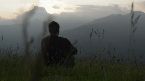 Young man seated on the grass is watching at panorama at sunset on mountain - HD video footage 库存视频