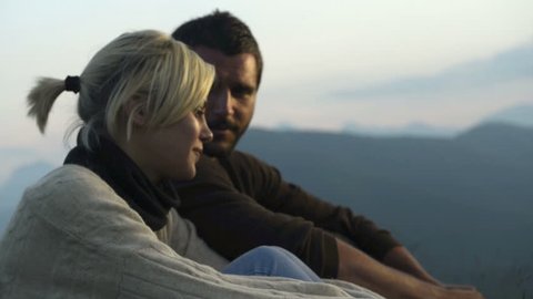Young beautiful romantic couple seated on the grass is watching at panorama at sunset on mountain - HD video footage, videoclip de stoc