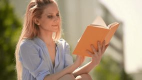 Young beautiful blonde girl in summer day is reading a book sit on a bech at town park - HD video footage