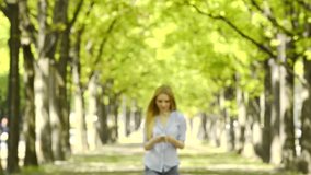 Young beautiful blonde girl in summer day is walking at town park listening music with smartphone - HD video footage