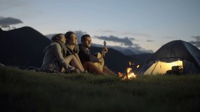 Group of three friends play guitar and sing at camp-fire in nature mountain outdoor camping scene at night - HD video footage