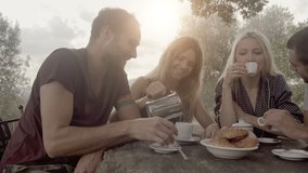 Group of four happy men and women friends smile, laugh and drink coffee during italian breakfast on a summer sunny day morning in tuscany, italy with visible sun - slow-motion dolly HD video footage