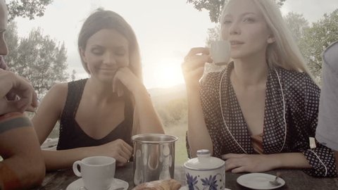 Group of four happy men and women friends smile, laugh and drink coffee during italian breakfast on a summer sunny day morning in tuscany, italy with visible sun - slow-motion dolly HD video footage 库存视频