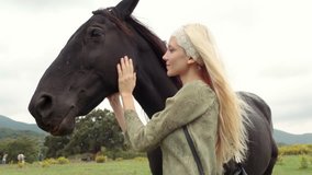 Blonde young woman smiles, strokes and hugs black horse outdoor and look at camera - HD video footage