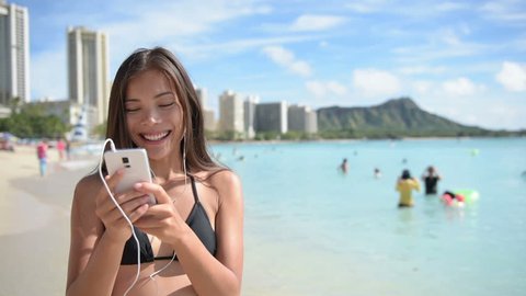 Woman using mobile cell smart phone laughing on beach wearing earphones for music or talking Girl in bikini using smartphone happy. Beautiful Asian Caucasian female model on Waikiki, Oahu, Hawaii, USA