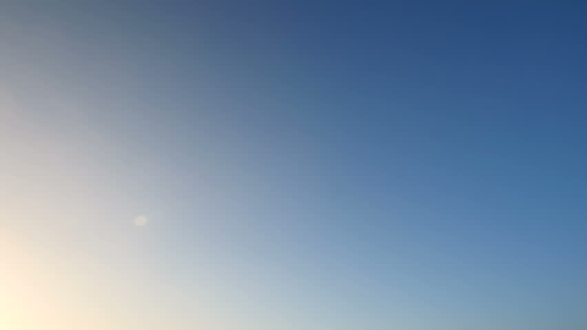 Airplane with red engines enters the frame overhead and flies away from the camera towards sunlight in a blue sky. Slow motion.