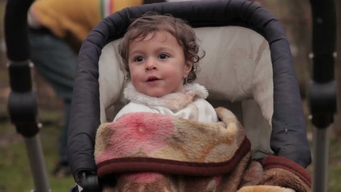 Profile of gypsy baby with beautiful smile. Little child ( kid ) in stroller covered with a blanket looking around. Facial expressions. Blurred people working in the background. Cute baby close up.
