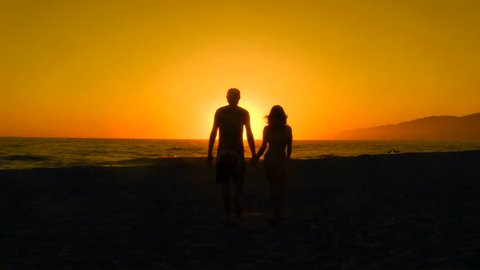 Romantic couple walks towards sunset holding hands 