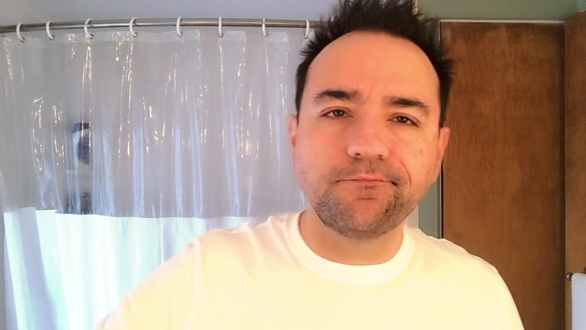 A tired and groggy man messily brushes his teeth in the morning while looking at himself in the bathroom mirror.