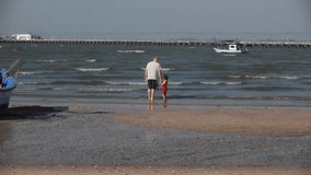 Stock Video Footage 1920x1080 1080p HDV Grandfather and granddaughter, family, goes to the coastal beach, goes to sea holding hands, sea, on the background of the pier, boat, nature, summer, vacation. 