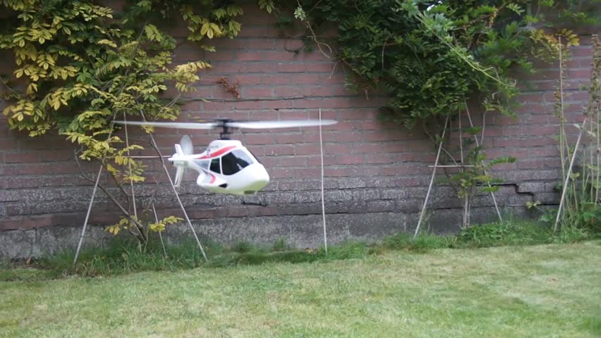 White small toy drone RC helicopter RC heli fly past low to the ground and close to the camera green grass and a wall at the background of footage RC pilot having trouble flying the aircraft steady 