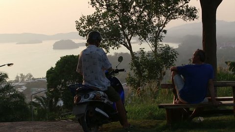 young guy in glasses and helmet sitting on scooter says goodbye to the elderly man sitting on bench under big tree and leaves