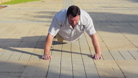 50 year old fat man tries to do push-ups on a pavement stone in summer day. But his efforts are unsuccessful
