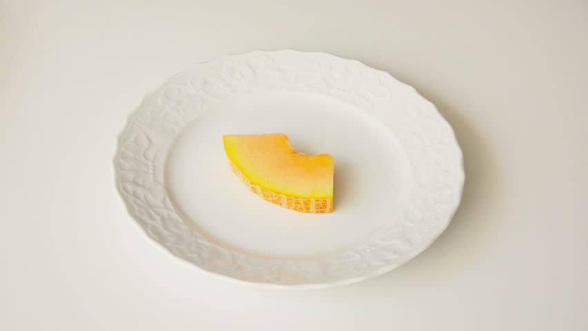 Cutting off a slice of melon with knife and fork on a white plate. Top view