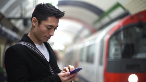 Young Asian man using his phone in a subway station as his train arrives