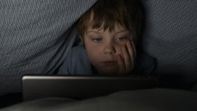 Young boy secretly plays on tablet at night under covers, past bedtime. 