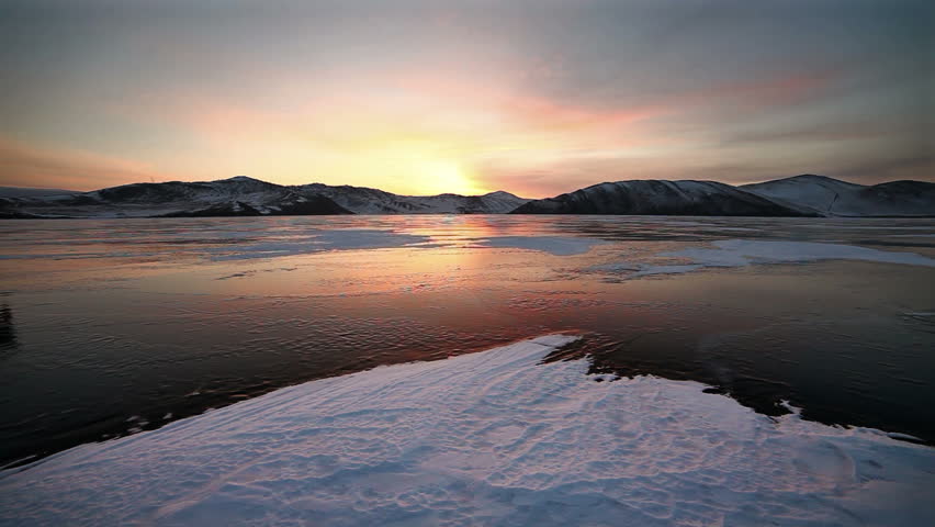 Tree backpackers walk on ice of lake against colorful sunset.