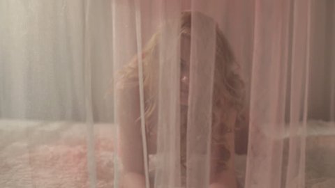 Curly blonde with long hair in lingerie kneeling on the bed behind transparent curtain