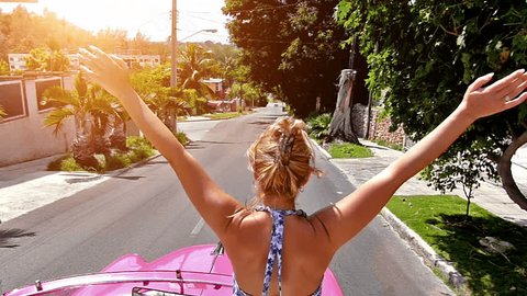 Rear view of happy blonde woman with arms outstretched, driving in pink cabriolet through Havana city, Cuba.Slow motion, high speed camera, lens flare
