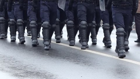 Heavily armored policemen with shields, bats, boots walking in disclpine on a riot public intervention.