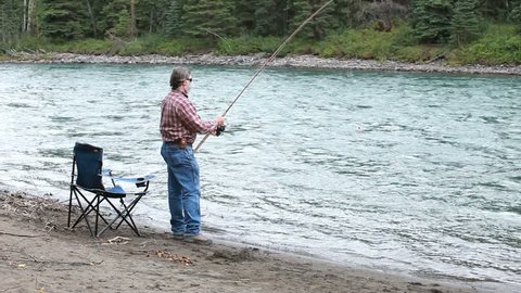 Man standing then sitting in chair on edge of river fishing. Beautiful blue green water. Sounds of water and birds chirping. Salmon run in Alaska and Canada.
