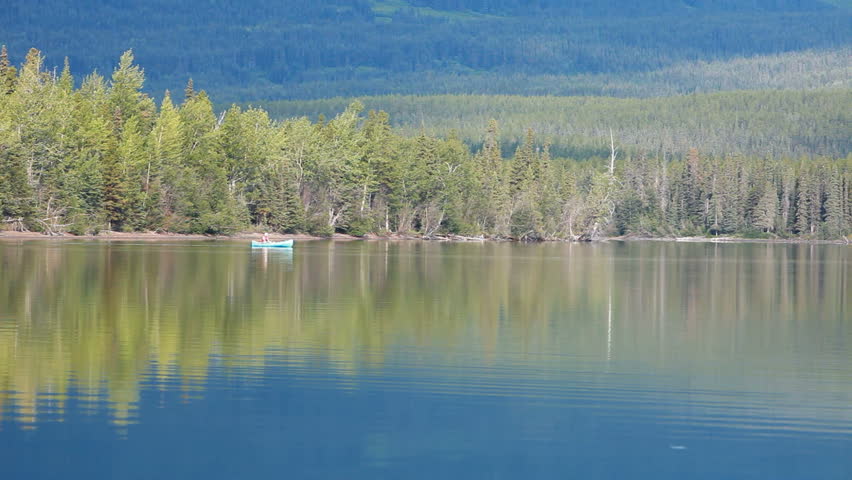 Canoe slowly paddled across beautiful calm mountain forest lake in British Columbia, Canada. Reflection of forest across lake. Vacation and recreation destination.