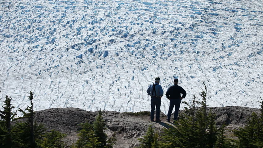 Men standing on mountain looking at Salmon Glacier in the distance. Pine forest contrasts with the blue glacial ice. Near Hyder British Columbia.