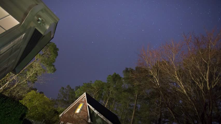 Time lapse video stars moving as camera pans across with trees and light pollution. Many planes fly streaking across night sky. House and roof can be seen.
