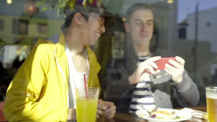 Multi-Ethnic Gay Couple Take A Selfie Together At Lunch (Shot From Outside, Looking In Through Window)
