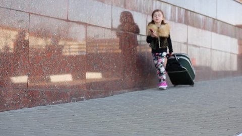 Pretty little girl dials a phone number and walking with suitcase on street.
