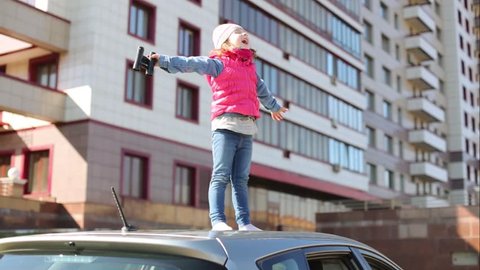 Little girl is standing on car roof with open hands.
