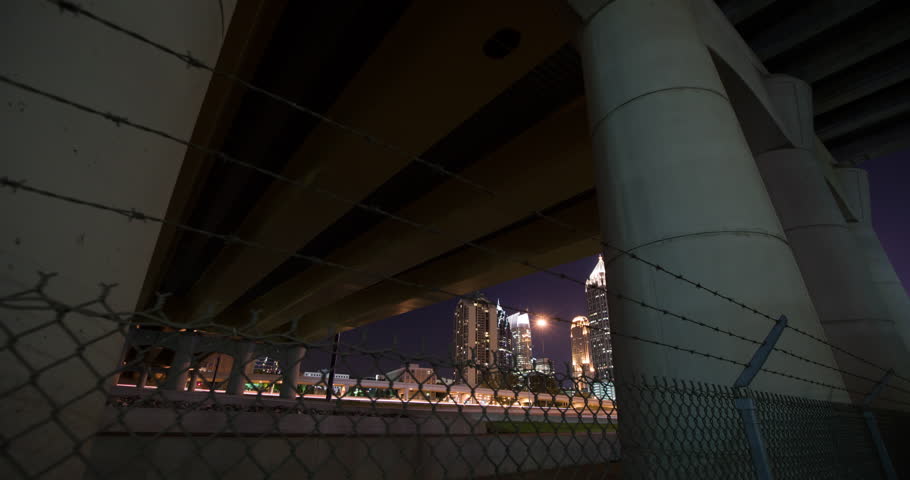 Timelapse of downtown Atlanta form under bridge by highway from behind a fence.  Symphony Tower, One Atlantic Center and Sun Trust Plaza in shot at night.