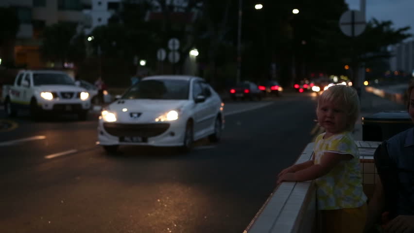 little blonde daughter and her mother sit on bench at pavement near city road and look at passing cars