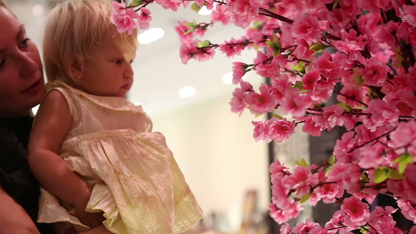 blonde child touches pink artificial sakura flowers sitting in mother arms in trade mall
